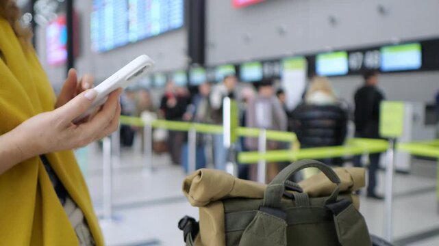 Woman in yellow jacket holds phone. Green backpack rests beside her. Airport check-in area with blurred travelers. Screens display flight information. Yellow stanchions mark queue lines