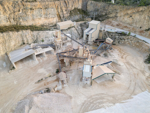 Aerial view of an industrial stone quarry with processing machinery, conveyor belts, and piles of gravel set against a rocky cliff face in Teulada, Valencian Community, Spain.