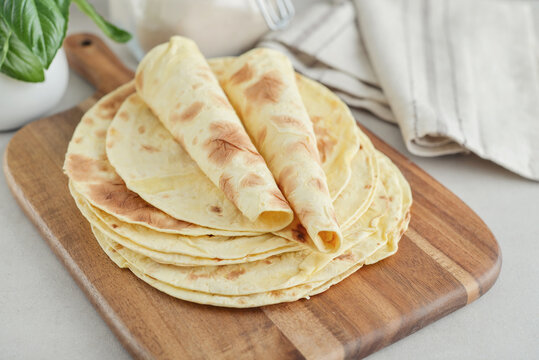 Stack of lavash or chapati flatbreads on a wooden cutting board, with a jar of flour, fresh basil
