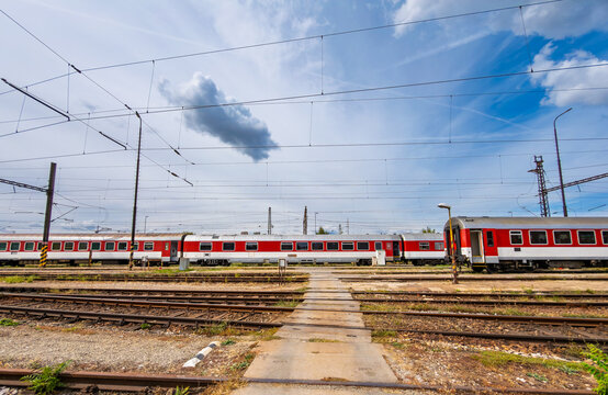 Red passenger train cars parked at railway station yard. Overhead power lines stretch above multiple tracks under a cloudy sky during daytime.