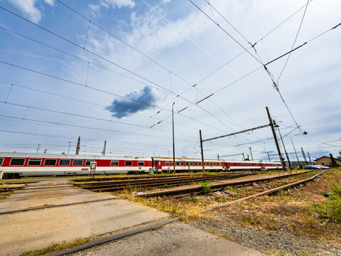 Parallel railway tracks and red passenger trains at depot. Wide angle view shows concrete ground crossing tracks beneath catenary lines during daytime.