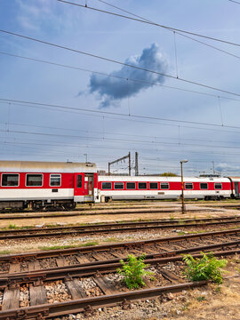Railroad tracks in foreground leading to passenger train. Steel railroad tracks with green weeds lead toward parked red train carriages under a clear blue sky.