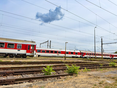 Railway tracks in foreground with parked passenger trains. Parallel steel railway tracks extend toward parked red train carriages under a blue sky with overhead lines.