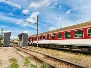 Passenger train carriage on railway tracks near a maintenance depot. A red and white rail car sits on steel tracks beside a metal service shelter under a blue cloudy sky. © Анна Терелюк