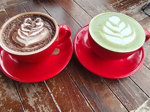 Red cups hold coffee and matcha art. Leaf designs float in creamy froths. Wood table adds rustic charm. Warm tones invite cozy moments. Perfect for cafe or wellness scenes