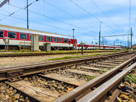 Curving railway tracks leading to passenger train carriages at a depot. Parallel steel rails curve across gray gravel ground towards red rail cars under a bright sky with light clouds.
