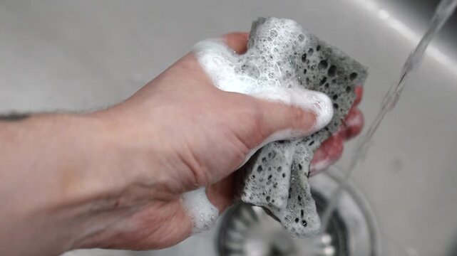 A man's hand squeezes water from a dish sponge over the kitchen sink.