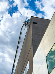 Aerial work platform extended along modern office facade. A hydraulic boom reaches the upper roof border of a tall gray architecture structure on a sunny day. © Анна Терелюк