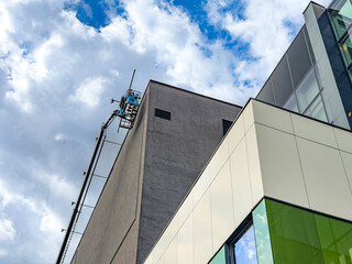 Worker in boom lift basket repairing high building facade. A technician in an aerial platform works near the top corner of a gray modern building under a cloudy sky. © Анна Терелюк
