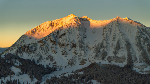 Aerial view of snow-covered peaks of the Ruby Range illuminated by golden sunrise light, with evergreen forests in the foreground near Crested Butte, Colorado, United States.