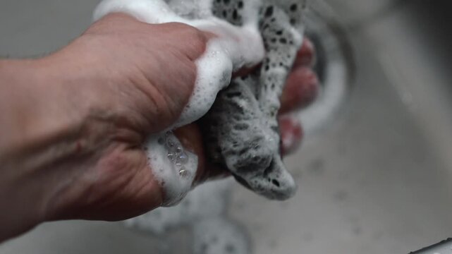 A man's hand squeezes water from a dish sponge over the kitchen sink.