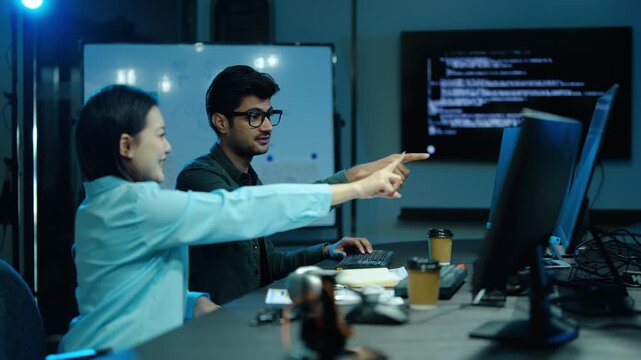 Diverse team of software developers working late in a modern tech office, programming and collaborating on code together, discussing project details on dual computer monitors in the dark room.