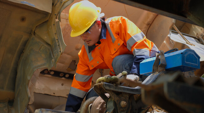 Professional mechanic in orange high visibility workwear performs hands maintenance inside an open heavy equipment engine compartment, showing industrial repair, technical skill and machinery service.