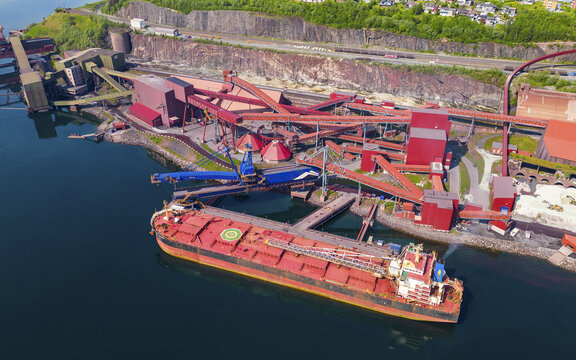 Aerial view of the industrial Narvik Harbor with a large red bulk carrier ship docked at the terminal, conveyor systems, storage silos, and rocky cliffs Narvik, Nordland, Norway.