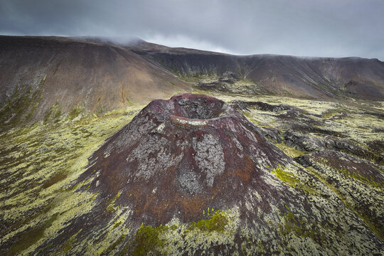 Aerial view of the volcanic cone surrounded by vast moss-covered lava fields under a dramatic cloudy sky, Eldborg Crater, Borgarbyggd, Iceland.