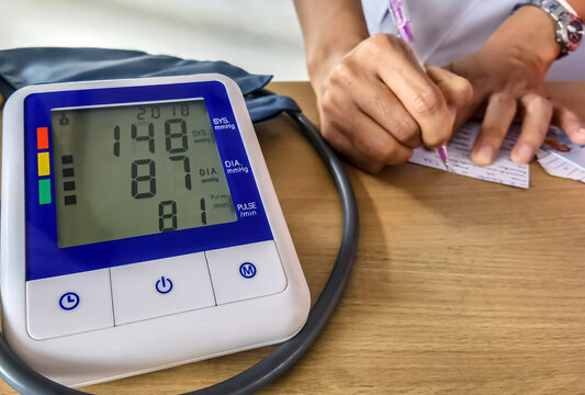Digital blood pressure monitor displaying blood pressure and pulse readings on wooden table with nurse using pen to record medical health check, blood pressure and pulse rate results in background.