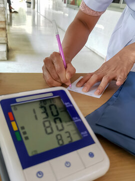 Hands of nurse using pen to writing blood pressure and pulse rate results record medical health from digital blood pressure monitor showing blood pressure and pulse rate readings on wooden table.