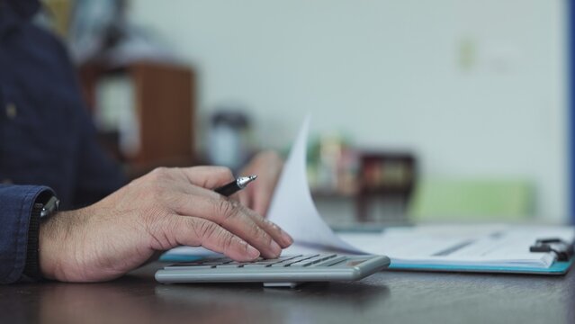 Businessman calculating financial paperwork beside clipboard on desk, analyzing accounting data, balance sheet figures, tax reports, business expenses.