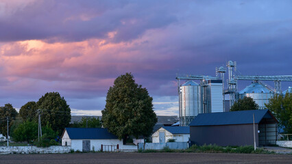 Rural agricultural landscape with grain silos and farm buildings under a dramatic purple and blue sunset sky. © kyrychukvitaliy