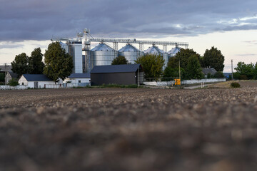 Agricultural grain storage facility with metal silos behind a plowed field in the countryside at dusk. Selective focus. © kyrychukvitaliy