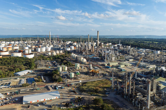 Aerial view of Oil Refinery with industrial storage tanks, tall chimneys, processing units, and surrounding greenery under a clear blue sky United Kingdom.