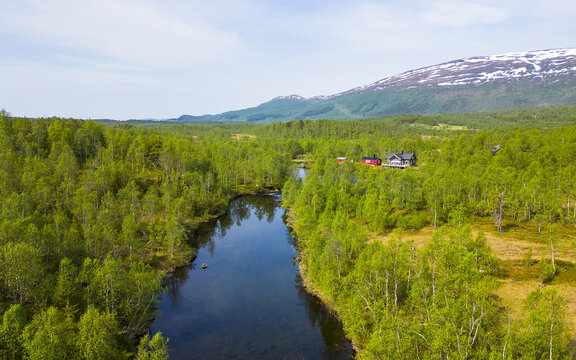 Aerial view of a river winding through a lush green forest with a small cabin in the background and snow-capped mountains under a clear sky in Narvik, Nordland, Norway.