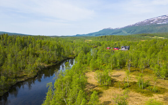 Aerial view of a river winding through a lush green forest with a small cabin nestled among trees and mountains in the distance in Narvik, Nordland, Norway.