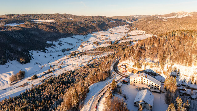 Aerial view of Col de la Faucille in winter, featuring snow-covered mountains, pine forests, a winding road, and buildings under golden sunlight in Gex, Auvergne-Rhone-Alpes, France.