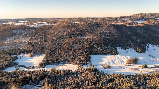 Aerial view of Col de la Faucille featuring snow-covered pine forests and open fields under clear winter light in Gex, Auvergne-Rhone-Alpes, France.