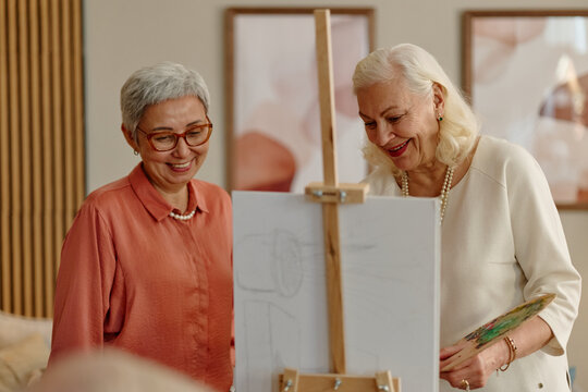 Senior Asian woman and senior Caucasian woman painting together on canvas during art workshop class. Scene represents active aging and creative recreation for retirement community campaigns