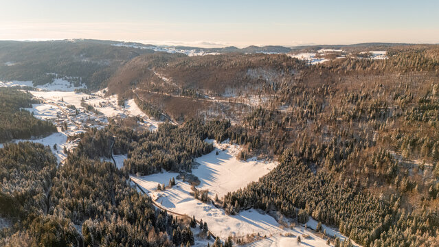 Aerial view of Col de la Faucille in winter, featuring snow-covered pine forests, rolling hills, and a small village nestled in the valley in Gex, Auvergne-Rhone-Alpes, France.