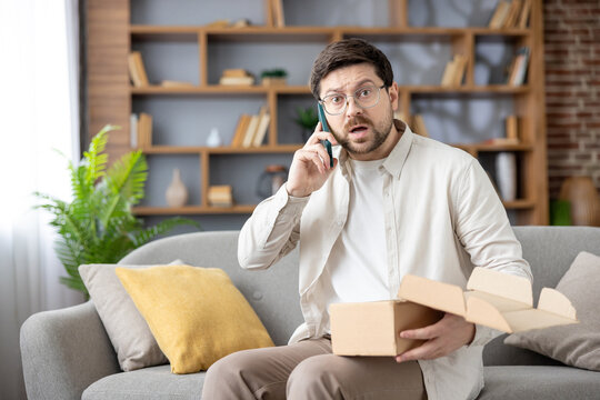 Man with a shocked expression holding an open, empty, or damaged delivery box while talking on a mobile phone, experiencing a customer service issue with his online order