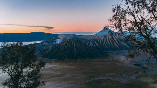 Aerial view of Mount Bromo with its dramatic volcanic crater, rugged slopes, and surrounding sea of sand under a soft sunrise sky in Tosari, East Java, Indonesia.