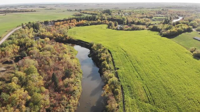 Drone flying over a green farm field and river surrounded by early fall colors
