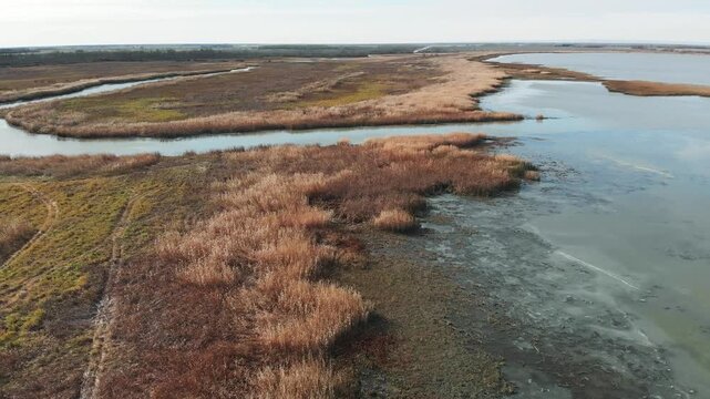 Drone flying low above a dry brown grassy shoreline and a small river with ice covered shallow water 
