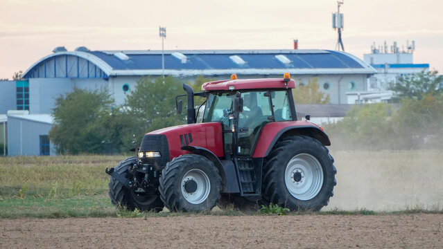 modern red tractor driving across an arable field raising a dust cloud in front of industrial buildings
