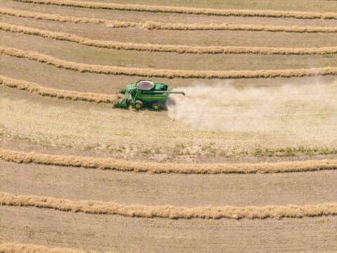 Aerial view of a green combine harvester working in a harvested wheat field, kicking up dust while moving through circular rows in Philadelphia, Western Cape, South Africa.