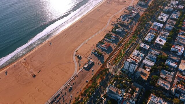 Lively road with multiple moving cars separates the cityscape from the beautiful empty sandy beach. Aerial view on the ocean shore in Los Angeles, California, USA lit by the bright sun.