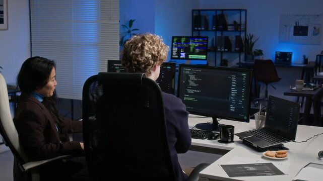 Young adult Asian woman and Caucasian man discussing software code on multiple monitors in office. Video ideal for cybersecurity training and software development campaigns