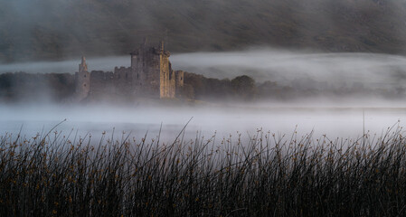 Dark reeds fill the foreground as low mist drifts across Loch Awe in front of the ruins of Kilchurn Castle, lit by soft early light. © Scott