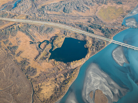 Aerial view of Knik River, showing the winding riverbed with glacial silt, a small dark lake surrounded by autumn trees, and a highway bridge in Knik River, Alaska, United States.
