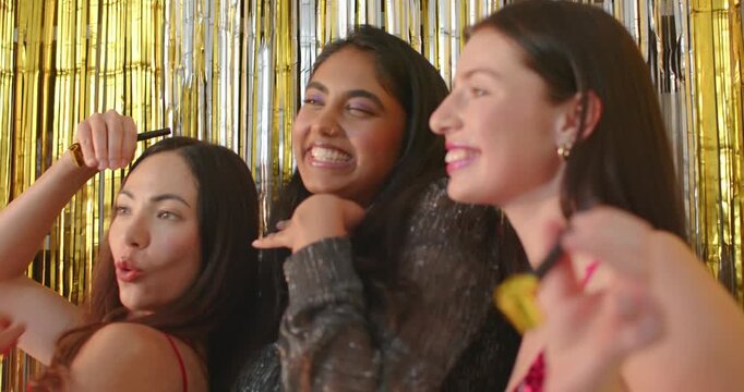 Diverse female friends posing at party booth, blowing gold horn, laughing under warm tinsel curtain