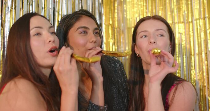 Diverse female friends blowing gold party blowers at foil backdrop after photobooth cue
