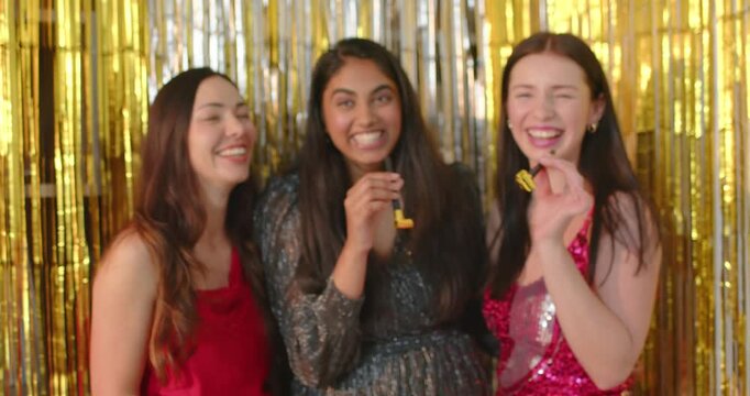 Three women laughing, trying and blowing gold horns in sequin dresses before gold tinsel