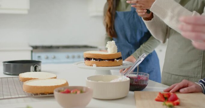 Hands are scooping icing, smoothing and slicing strawberries, decorating cake on kitchen counter