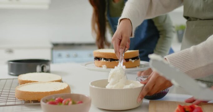 Diverse female friends wearing aprons scooping, smoothing frosting on cake for dessert in kitchen