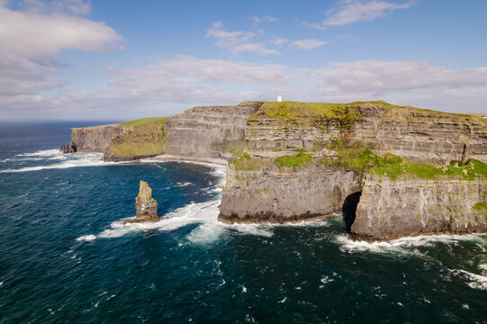 Aerial view of the Cliffs of Moher with rugged rock faces, green grassy clifftops, and crashing waves in the Atlantic Ocean in Ballysteen, Clare, Ireland.