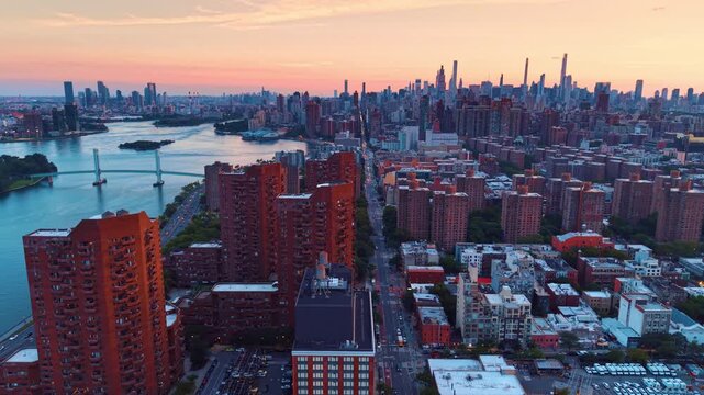 Moving over high-rise red brick buildings at the Harlem River waterfront. Approaching the Wards Island Bridge across the river and Manhattan skyline against pink sly at sunset.