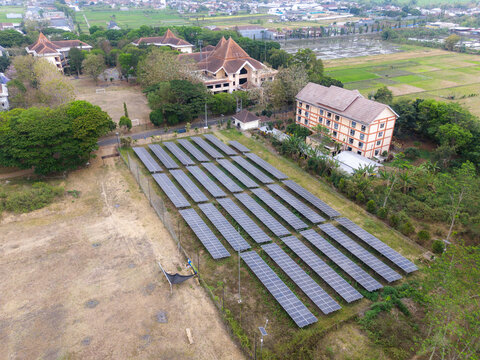 Aerial view of a solar panel array installed in a field near buildings and lush trees in Indonesia.