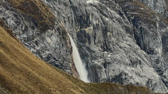 Powerful waterfall plunges down dramatic dark mountain cliff amidst golden ichu grass slope near tres lagunas sector in cordillera huayhuash peruvian andes
